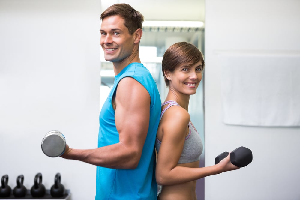 Fit couple lifting dumbbells together smiling at camera at the gym